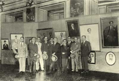 A fotografia está em preto e branco e mostra um grupo de pessoas em frente a vários quadros. A maioria é de homens, que estão em pé e posando para o fotógrafo. Eles vestem terno e seguram seus chapéus à frente do corpo. Estão em um lugar fechado e amplo e, na parede atrás deles, há vários quadros de tamanhos diferentes. 