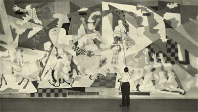 Foto, em preto e branco, de um homem em frente a um painel gigante. Ele está em pé, de costas e olhando para cima. O painel, que tem aproximadamente três vezes a altura do homem, integra o Monumento Rodoviário, entre Rio de Janeiro e São Paulo. As imagens pintadas na imensa parede são um aglomerado de diferentes figuras e formas.