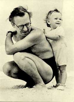 Fotografia, em preto e branco, de um homem e uma criança. Trata-se do artista e seu único filho, João Cândido. Aparecem em close, de perfil esquerdo sobre uma porção de areia. O homem está agachado, olhando para o chão e vestindo somente uma sunga, além de óculos. A criança veste roupa clara e está em pé, de frente para as costas do homem, onde apoia as mãos.