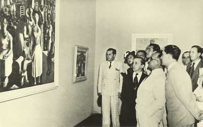 Em preto e branco, a fotografia revela um grupo observando um grande quadro. Estão em pé, acumulados na lateral direita. O quadro para o qual olham retrata pessoas. Na mesma parede, e na parede ao fundo, há dois quadros menores.