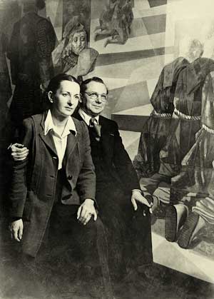 Foto em preto e branco de um casal em frente a uma das obras do artista, chamada “A primeira missa”. Os dois estão na diagonal, quase de frente para a câmera. A mulher está bastante próxima à esquerda e é abraçada por um homem. Somente o braço direito dele a envolve, passando pelas costas, e terminando na mão, que segura o braço dela. A tela é enorme e ocupou todo o espaço da imagem.