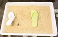 Sobre uma porção de areia batida, um chinelo verde é colocado em cima da superfície e deixa marca uniforme do seu solado na areia. Em seguida, uma sandália branca de salto faz o mesmo movimento, mas a marca não é constante como na primeira pegada, pois deixa um buraco na areia, exatamente onde houve a pressão do salto.