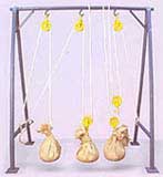 A ilustração colorida apresenta um suporte com um sistema de roldanas. O suporte está fixado ao chão e o sistema montado inclui três roldanas na parte de cima e três na parte de baixo, por onde passam cordas. Essas estão amarradas nas laterais inferiores do suporte e sustentam três sacos.