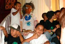 Nesta fotografia em cores, veem-se, em close, algumas crianças. Nota-se que estão sentadas nos degraus de uma escada. Percebe-se ainda que três meninos estão de frente para a câmera e um deles faz o sinal de positivo. Atrás desses, há um boneco, além de outras crianças olhando para a direita. 