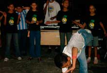 Nesta foto em cores e em close, um menino está com uma das mãos no chão e uma perna para o alto. Atrás dele, há outros pré-adolescentes em pé, vestindo camisetas pretas. Um deles usa camisa de futebol. Ao fundo, atrás do grupo, nota-se ainda um homem às costas de uma mesa com equipamentos de som.