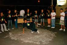 Um grupo de pré-adolescentes está em pé, de frente para a câmera e lado a lado, formando uma meia lua. Nota-se que um deles se destaca por estar à frente aos demais. Esse menino movimenta-se colocando uma das mãos no chão, apoiando o corpo, que quase deita no piso. Ao fundo, observa-se um homem às costas de uma mesa.
