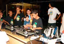 Esta foto colorida mostra, em close, alguns garotos ao redor de uma mesa de som utilizada por DJ's. Um deles manuseia o LP e os botões do equipamento, enquanto os demais apenas o observam. Nota-se que há um homem em pé, ao fundo da imagem.