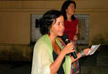 Nesta fotografia em cores e em ângulo fechado, uma mulher destaca-se por estar no centro, falando ao microfone. Nota-se que ela segura um pedaço de papel próximo ao rosto, mas olha para frente. Ao fundo nota-se a presença de outra mulher.