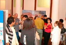 Nesta fotografia colorida, várias pessoas estão juntas em um salão onde há painéis da mostra. Os visitantes movimentam-se em diferentes direções, alguns conversam em pequenos grupos. Observam-se os portais de acesso a outro ambiente, além de colunas redondas igualmente altas.