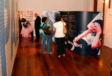 Em um salão, veem-se algumas pessoas observando painéis da exposição. A Foto, tirada em perspectiva, mostra um grande painel à esquerda, que toma a parede inteira e onde é possível ler a palavra 'PAZ' em letras gigantes. 