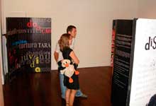 Nesta foto, um homem e uma mulher jovens apreciam painel exposto em um salão. O casal ocupa o centro da imagem e observa a obra à direita. Ao fundo, no lado esquerdo, nota-se um outro painel, com letras coloridas que se destacam sobre fundo preto. 