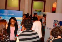 Nesta fotografia colorida, várias pessoas estão juntas em um salão onde há painéis da mostra. Observa-se que os visitantes estão de costas para a câmera, além de movimentarem-se em diferentes direções ou conversarem em pequenos grupos. Notam-se também os portais de acesso a outro ambiente, além de colunas redondas igualmente altas.